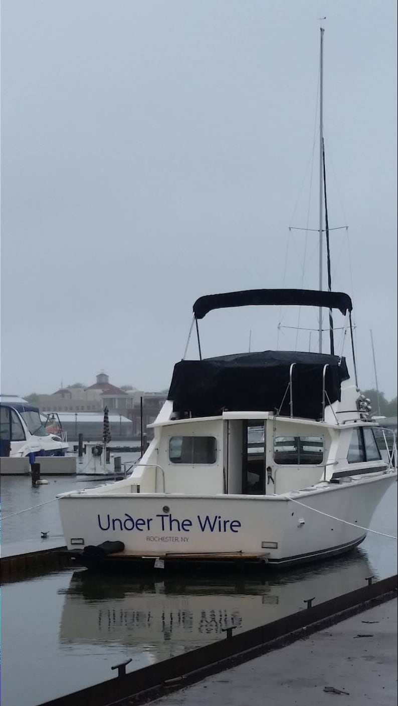 K&ouml;nnte beinhalten: Ein wei&szlig;es Boot, das an einem Steg in einer Marina liegt, mit einem schwarzen Stoffverdeck und dem Schriftzug "Under The Wire Rochester, NY" an der Seite.