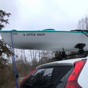 K&ouml;nnte beinhalten: Ein wei&szlig;es Kajak mit t&uuml;rkisfarbenem Rand ist auf dem Dach eines wei&szlig;en Autos befestigt. Das Kajak hat einen schwarzen Aufkleber mit der Aufschrift "A LITTLE NAUTI".