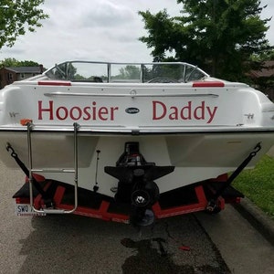 K&ouml;nnte beinhalten: Ein wei&szlig;es Boot mit der Aufschrift "Hoosier Daddy" in Rot auf der R&uuml;ckseite. Das Boot steht auf einem Anh&auml;nger mit einer Leiter an der Seite.