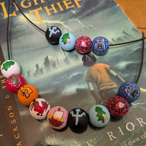May include: A necklace featuring a series of painted wooden beads with various designs, including a tree, a maze-like pattern, and other symbols. The necklace is displayed on a book titled "The Lightning Thief" by Rick Riordan.