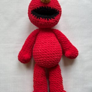 May include: A handmade red crochet doll resembling a popular children's character. The doll has a round head with white and black eyes, a brown nose, and a wide black mouth. It has arms, legs, and feet, all in the same vibrant red color.