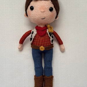 May include: A handmade crochet doll resembling a cowboy character. The doll features a brown hat, hair, and boots, with a red shirt, blue jeans, and a vest with black spots. A yellow sheriff's badge is pinned on the shirt.