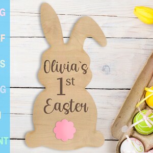 Puede incluir: Un recorte de madera de un conejo con el texto "Olivia's 1st Easter" y una flor rosa en la parte inferior. El conejo está sentado sobre una superficie de madera blanca.