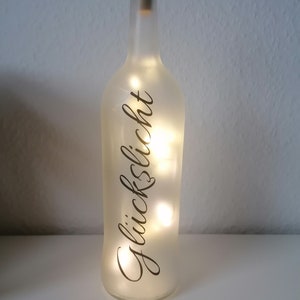 Könnte beinhalten: Eine gefrostete Glasflasche mit einem Korkverschluss und einer Lichterkette im Inneren. Die Flasche ist mit dem Wort "Glückslicht" in schwarzer Schrift verziert.