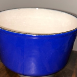 May include: A deep, round cooking pot with a vibrant cobalt blue exterior and a smooth, cream-colored interior. The pot features two integrated side handles and a dark, metallic-toned rim around the top edge, showcasing a sturdy construction.
