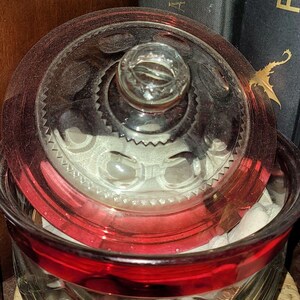 May include: A clear glass jar with a red rim and a matching glass lid. The jar features a diamond-cut pattern on the exterior, while the lid has a circular knob handle and a decorative scalloped edge. The interior contains white fabric items.