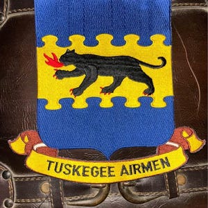 Puede incluir: Un parche de tela con un escudo azul y una banda horizontal amarilla. Una pantera negra con garras rojas y una llama roja en la boca está centrada en la banda amarilla. Debajo del escudo hay una cinta amarilla con el texto TUSKEGEE AIRMEN en negro.