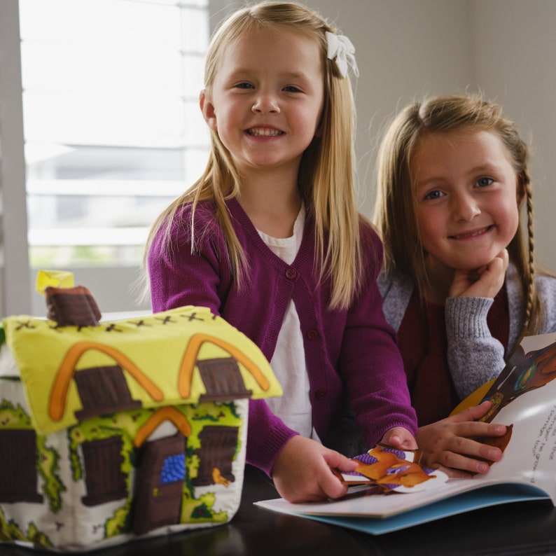 May include: Two young girls smiling, one holding a colourful storybook and the other a fabric house. The house has a yellow roof, brown accents, and green and white walls. The girls are wearing purple and grey cardigans.