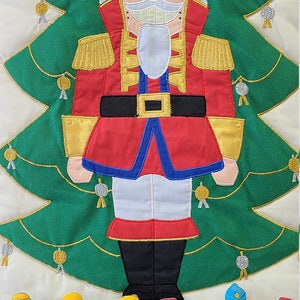 May include: A green felt advent calendar with a Nutcracker theme. The calendar features a large embroidered Nutcracker figure standing in front of a green Christmas tree. There are 13 pockets on the calendar, each with a different character from the Nutcracker story. The pockets are numbered 1 through 13.
