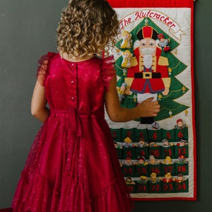 May include: A girl wearing a red sparkly dress is looking at a felt advent calendar with a Nutcracker theme. The calendar has 24 pockets with numbers 1-24 and a Nutcracker figure in the centre. The text "The Nutcracker" is at the top of the calendar.