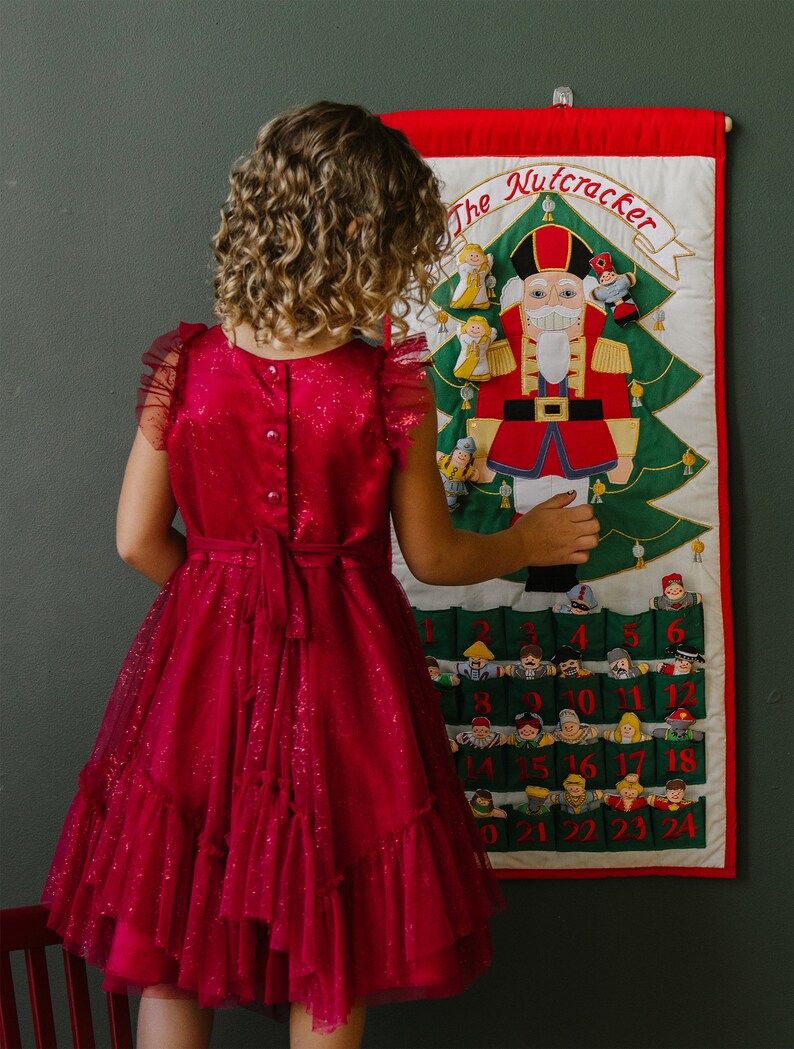 May include: A girl wearing a red sparkly dress is looking at a felt advent calendar with a Nutcracker theme. The calendar has 24 pockets with numbers 1-24 and a Nutcracker figure in the centre. The text "The Nutcracker" is at the top of the calendar.