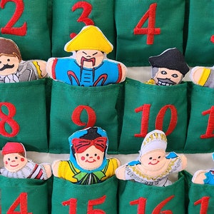 May include: A green felt advent calendar with 18 pockets, each featuring a different embroidered character and a red number. The characters are dressed in various costumes, including a pirate, a king, and a clown.