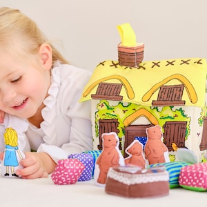May include: A child's toy set featuring a fabric house with a yellow thatched roof, brown windows, and a chimney. The set includes bear figures, a girl figure in a blue dress, and colourful fabric cushions. The child is smiling.