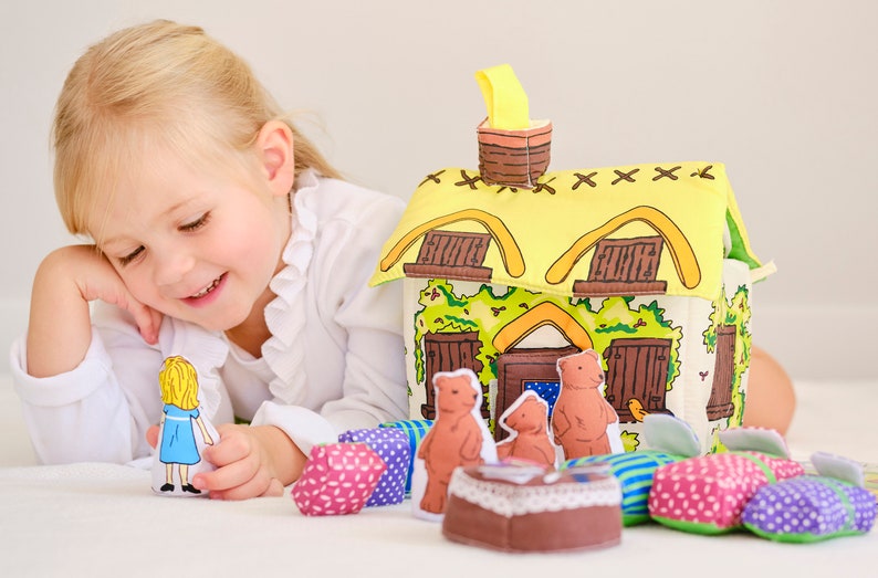 May include: A child's toy set featuring a fabric house with a yellow thatched roof, brown windows, and a chimney. The set includes bear figures, a girl figure in a blue dress, and colourful fabric cushions. The child is smiling.