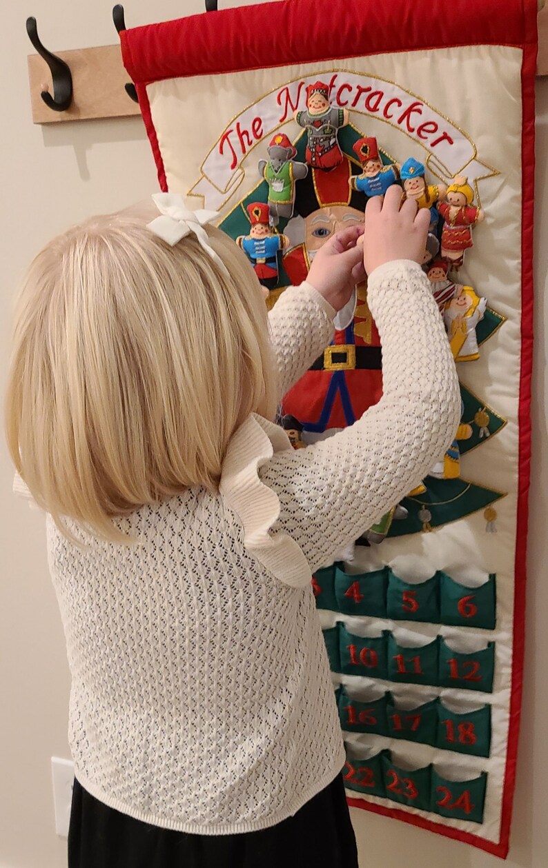 May include: A young child wearing a white knitted sweater and a black skirt is interacting with a felt advent calendar featuring a Nutcracker theme. The calendar has 24 pockets with numbers 1 through 24. The child is opening a pocket on the calendar.