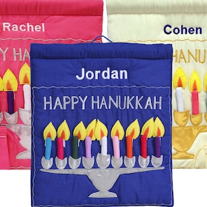 May include: A blue fabric Hanukkah banner with the name Jordan embroidered on it. The banner features a menorah with nine candles and the text "Happy Hanukkah".
