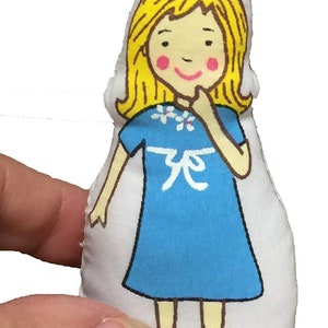 May include: A handmade fabric doll featuring a cartoon girl illustration. The doll has yellow hair, pink cheeks, and wears a blue dress with a white floral design and a white bow. The doll is approximately 10 cm tall.