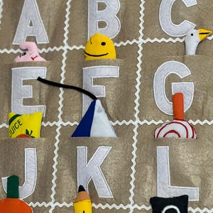 May include: A felt alphabet chart with pockets for small toys. Each letter is white and outlined in white. The pockets contain a variety of small toys, including a book, a pencil, a juice box, a pumpkin, a smiley face, a bunny, a goose, a cone, and a swirl.