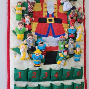May include: A green and white fabric advent calendar with 24 pockets. The calendar features a Nutcracker theme with embroidered characters and a large Nutcracker figure in the centre. The numbers 1 through 24 are embroidered on the pockets.