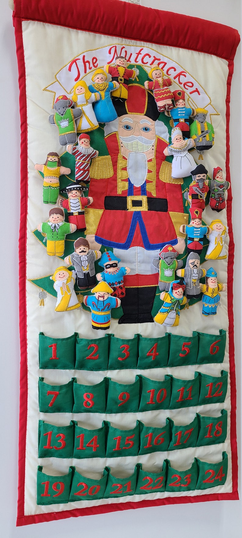May include: A green and white fabric advent calendar with 24 pockets. The calendar features a Nutcracker theme with embroidered characters and a large Nutcracker figure in the centre. The numbers 1 through 24 are embroidered on the pockets.
