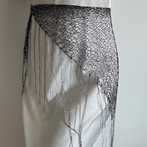May include: A black, open-weave belt with a triangular design and dangling fringe. The belt is worn over a white garment, creating a contrast in textures and colors. The fringe hangs down, adding movement to the design.