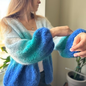 May include: A blue, green, and white ombre cardigan with a fuzzy texture. The cardigan has a V-neck and long sleeves.