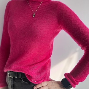 Op de afbeelding: Een felroze coltrui met lange mouwen. De trui wordt gecombineerd met een zwarte riem en een grijze jeans. Een zilveren kruis-ketting is zichtbaar. De mouwen zijn bij de polsen opgerold.