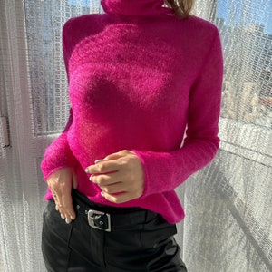 Op de afbeelding: Een fel fuchsia coltrui met lange mouwen. De trui wordt gecombineerd met een zwarte leren broek met een zilveren gesp. De outfit staat voor een raam met zonlicht.