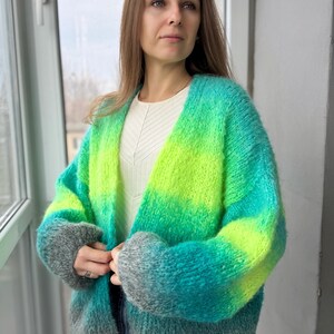 May include: A green, blue, and gray ombre cardigan with a loose fit. The cardigan has a V-neck and long sleeves. The word "SEA" is printed on the bottom right corner of the image.
