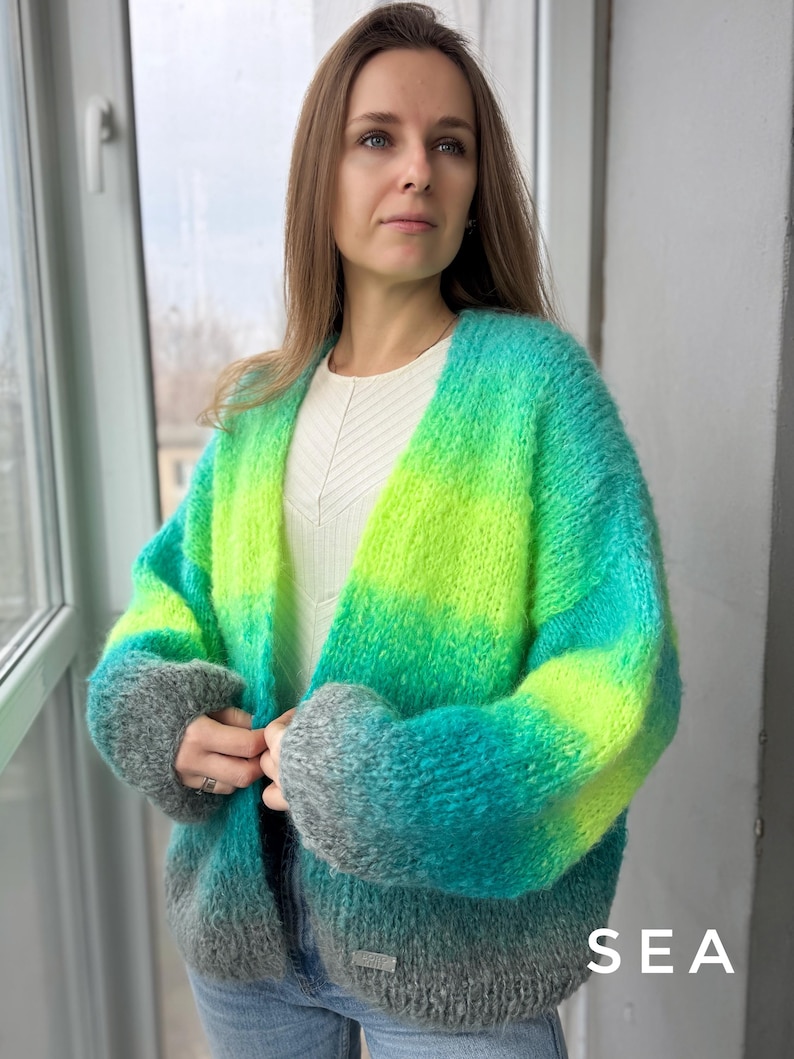 May include: A green, blue, and gray ombre cardigan with a loose fit. The cardigan has a V-neck and long sleeves. The word "SEA" is printed on the bottom right corner of the image.