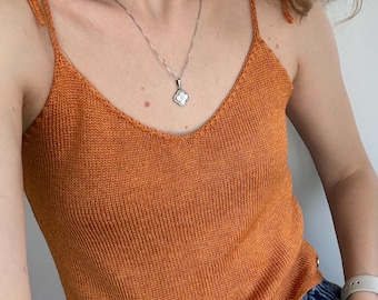 May include: An orange knitted camisole top with tie straps. The top has a V-neck and a ribbed texture. A silver necklace with a clover pendant is worn with the top. The top is paired with blue denim jeans.