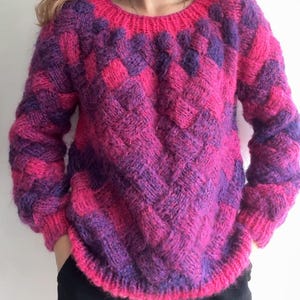 May include: A hand-knitted jumper in shades of pink and purple. The jumper features a crew neck and long sleeves. The design incorporates a textured, geometric pattern. The garment is made of a soft, fuzzy yarn.