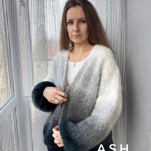 May include: A gray and green ombre cardigan with a V-neck and long sleeves. The cardigan is open in the front and the sleeves are rolled up. The model is wearing black pants and a silver necklace. The word "ASH" is printed on the bottom of the cardigan.