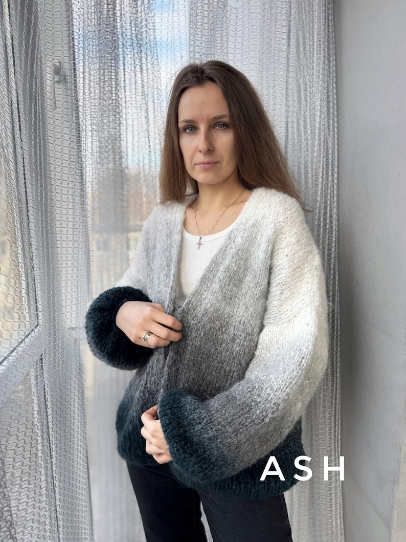 May include: A gray and green ombre cardigan with a V-neck and long sleeves. The cardigan is open in the front and the sleeves are rolled up. The model is wearing black pants and a silver necklace. The word "ASH" is printed on the bottom of the cardigan.