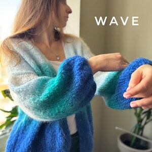 Puede incluir: Un c&aacute;rdigan azul, verde y blanco degradado con una textura esponjosa. El c&aacute;rdigan tiene cuello en V y mangas largas. La palabra "WAVE" est&aacute; escrita en blanco sobre un fondo blanco.