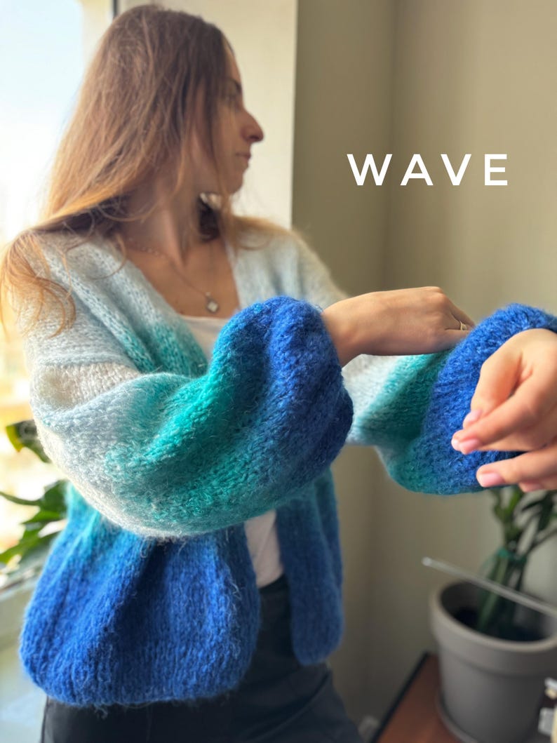 Puede incluir: Un c&aacute;rdigan azul, verde y blanco degradado con una textura esponjosa. El c&aacute;rdigan tiene cuello en V y mangas largas. La palabra "WAVE" est&aacute; escrita en blanco sobre un fondo blanco.