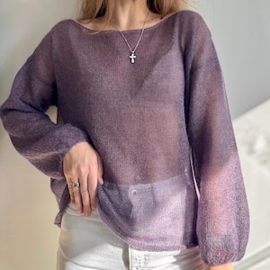 May include: A sheer, long-sleeved purple sweater with a boat neck. The sweater has a loose, flowing fit and is made of a lightweight, possibly knitted material. The sleeves are gathered at the wrist. A silver cross pendant hangs from a delicate chain.