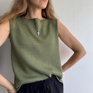 May include: Olive green sleeveless top with a textured knit design. The top is paired with black drawstring pants. A silver cross pendant hangs from a delicate chain. The model is wearing a silver bracelet.