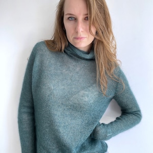 Pu&ograve; includere: Un maglione a maniche lunghe color verde acqua con collo alto. Il capo &egrave; realizzato in un tessuto a maglia fine e semitrasparente con una trama visibile. Il maglione &egrave; abbinato a un paio di jeans in denim grigio chiaro con cintura.