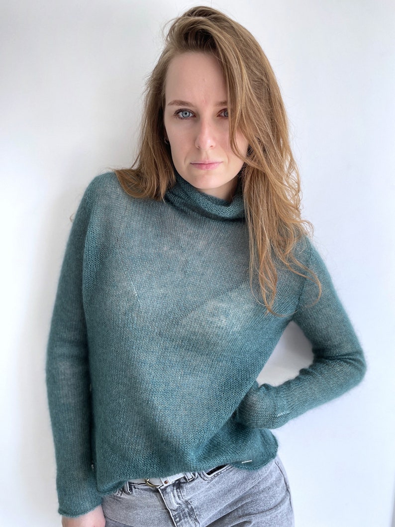 Pu&ograve; includere: Un maglione a maniche lunghe color verde acqua con collo alto. Il capo &egrave; realizzato in un tessuto a maglia fine e semitrasparente con una trama visibile. Il maglione &egrave; abbinato a un paio di jeans in denim grigio chiaro con cintura.