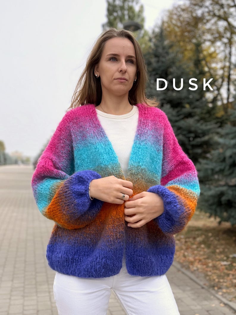 Puede incluir: Un c&aacute;rdigan de color degradado con un degradado de rosa, azul, naranja y morado. El c&aacute;rdigan tiene cuello en V y mangas largas. La palabra "DUSK" est&aacute; escrita en blanco en el lado derecho de la imagen.