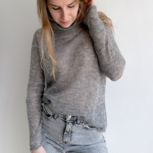 Pu&ograve; includere: Un maglione a collo alto grigio a maniche lunghe, semi-trasparente, abbinato a jeans in denim grigio chiaro. Il maglione presenta una trama fine e leggera con una vestibilit&agrave; ampia e rilassata che cade sul busto e sulle braccia.