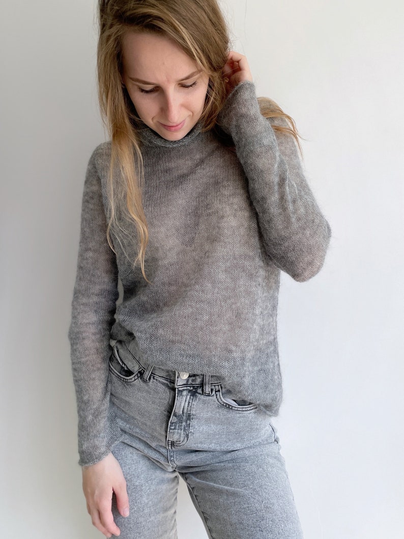 Pu&ograve; includere: Un maglione a collo alto grigio a maniche lunghe, semi-trasparente, abbinato a jeans in denim grigio chiaro. Il maglione presenta una trama fine e leggera con una vestibilit&agrave; ampia e rilassata che cade sul busto e sulle braccia.