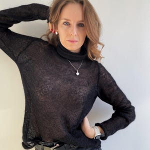 Può includere: Una donna indossa un maglione nero a collo alto trasparente con una collana d'argento e una cintura nera su un paio di jeans neri.