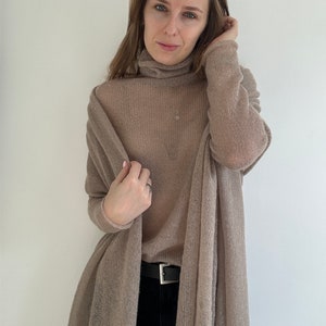 Pu&ograve; includere: Un maglione a collo alto beige, semi-trasparente, a maniche lunghe con una vestibilit&agrave; ampia e drappeggiata. Il capo &egrave; abbinato a una cintura nera con fibbia rettangolare color argento, indossata sopra pantaloni scuri. Il tessuto ha una trama fine.