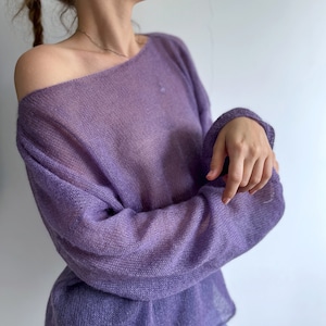 Puede incluir: Un suéter lavanda con hombros descubiertos. El tejido de punto tiene un tejido suelto, lo que le da un aspecto semitransparente. Las mangas largas están abrochadas en las muñecas. El suéter es de color liso y parece estar hecho de un material suave y ligero.