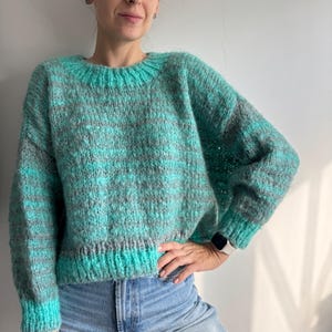 May include: A teal, grey, and turquoise knitted jumper. The jumper has a crew neck and ribbed cuffs and hem. The yarn has a textured appearance. The model is wearing blue jeans.