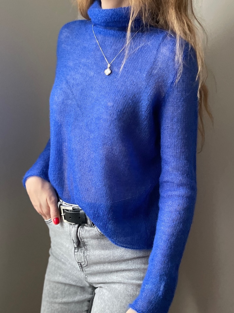 Pu&ograve; includere: Un maglione a maniche lunghe di colore blu reale vibrante con collo alto, caratterizzato da una trama a maglia leggera e trasparente. Il maglione &egrave; abbinato a jeans in denim grigio chiaro e a una cintura in pelle nera con fibbia argentata.