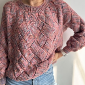 May include: A pink, red, and blue knitted jumper with a diamond pattern. The jumper has a crew neck and long sleeves. The sleeves have a puffy design. The jumper is paired with blue jeans.