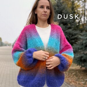 May include: A colorful ombre cardigan with a gradient of pink, blue, orange, and purple. The cardigan has a V-neck and long sleeves. The word "DUSK" is written in white text on the right side of the image.